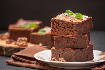Chocolate brownie square pieces in stack on white plate with walnuts, decorated with mint leaves and cocoa on black background. Delicious dessert. Dark mood. Close up photography. Selective focus