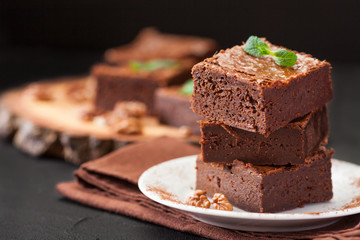 Chocolate brownie square pieces in stack on white plate with walnuts, decorated with mint leaves and cocoa on black background. Delicious dessert. Dark mood. Close up photography. Selective focus