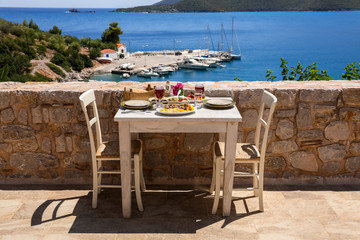 A table served for two with chicken souvlaki and french fries, greek salad, snacks and drinks on the summer terrace of the hotel room by the seascape, beautiful summer greek holidays concept.