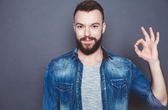 It`s good. A handsome smiling modern man in a denim shirt shows a hand sign ok on a gray background.