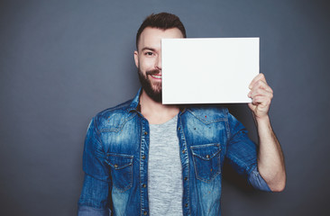 Everything is for you. Handsome young smiling man in a denim shirt shows a white sheet of paper in the camera on a gray background. Area for advertising.