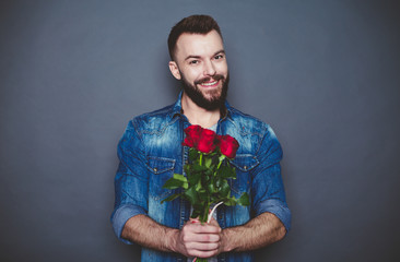 Gift for the beloved. Beautiful smiling bearded man in a denim shirt holds a red rose on a gray background.