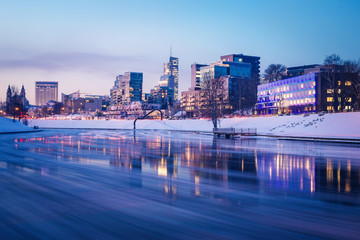 Cityscape skyline with office buildings on a river bank, clear blue sky, and reflections on the water during twilight blue hour in Vilnius, Lithuania