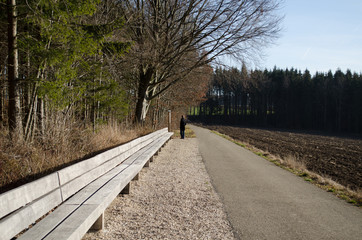 Lange Bank die ins Bild hineinführt mit Feldweg der parallel dazu verläuft und Mann vor einem Schild steht