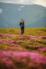 Naklejka na ściany i meble Shot of a young woman looking at the landscape while hiking in the mountains. Flowers in the mountains. Majestic flowers glowing by sunlight. Magic pink rhododendron flowers on summer mountain