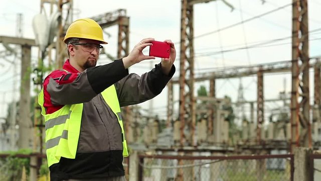 Worker records videos on smartphone. Powerman in yellow hard hat with smartphone at electric power plant