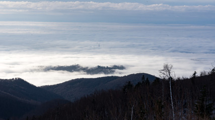 Fototapeta na wymiar Landscape view from Sljeme or Medvednica mountain down on clouds covering the city of Zagreb in Croatia with a visible smoking heat plant chimney above the clouds during sunrise