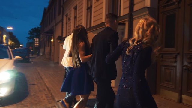 Group Of Young Party People With Champagne Walks On Night Street Road Toward House Doors In Front Of White Car Headlights