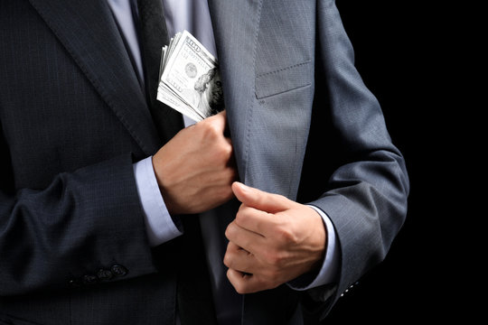 Businessman putting banknotes in pocket on dark background. Corruption concept