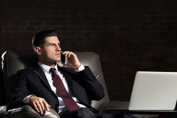 Handsome young man in suit talking by phone while sitting in armchair indoors