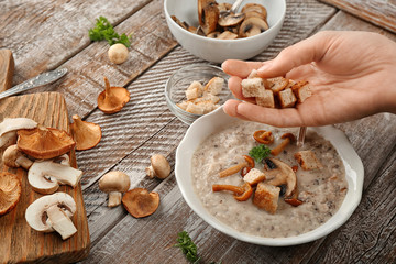 Young woman adding croutons to delicious mushroom soup at wooden table