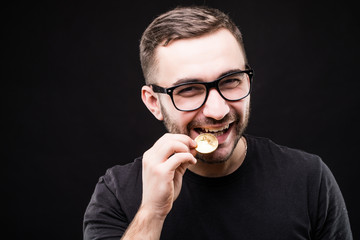 Close up portrait of a man in glasses biting golden bitcoin isolated over black background