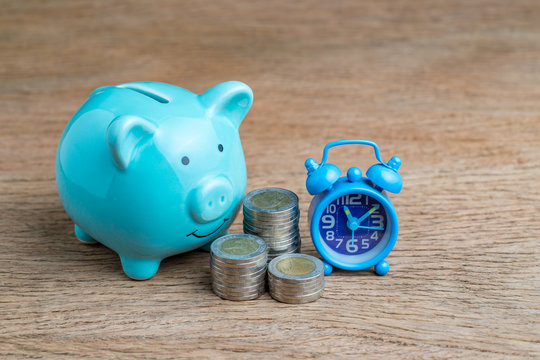 Financial long term saving money concept, blue piggy bank, coins stacked and alarm clock on wood table, compound interest of investment awareness
