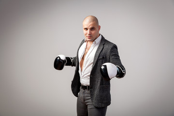 A young bald man in an unbuttoned white shirt, gray suit and boxing gloves looks defiantly into the camera and posing on a white isolated background