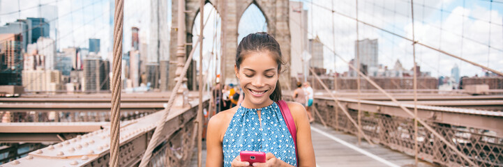 Happy woman using phone walking in NYC on Brooklyn Bridge. New York city lifestyle young Asian girl...