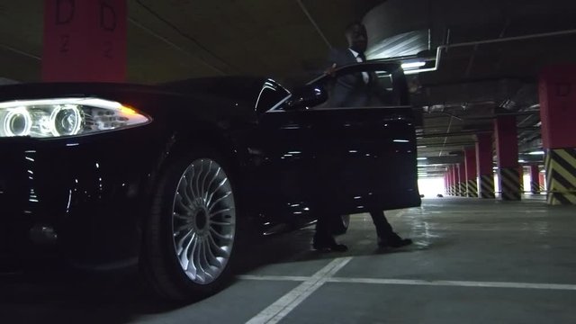 Pan with low angle of successful African businessman in suit getting out of his exPansive car in underground parking lot