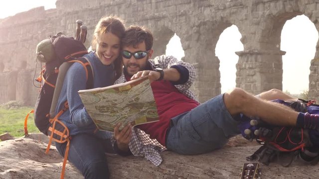 Happy young couple backpackers tourists sitting lying on a log trunk reading map guide pointing ancient roman aqueduct ruins in parco degli acquedotti park in rome at sunrise romantic with sleeping