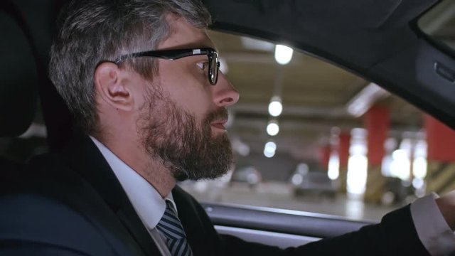 Medium shot of bearded businessman wearing suit and glasses driving car in underground parking lot
