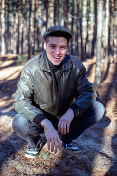 A guy in a leather jacket and cap sits on a road in a pine forest.