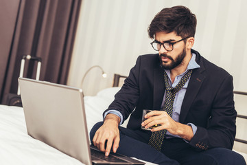 Working in hotel room. Confident young businessman in suit and tie working on laptop while sitting...