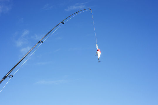The spinning and brightly-colored crankbait are against the blue cloudless sky. The fishing equipment is on a cyan background. It is cast a line.