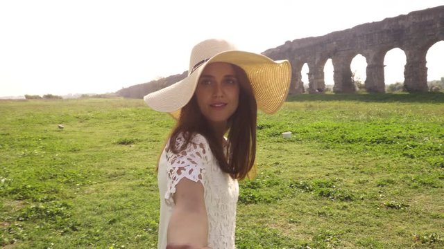 Come with me. Young beautiful girl with hat leading man boyfriend holding hands toward roman aqueduct arches in parco degli acquedotti park ruins in rome romantic lovely happy couple slow motion