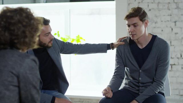 PAN of distress young man talking and sharing feelings as bearded male patient supporting him during interpersonal group psychotherapy