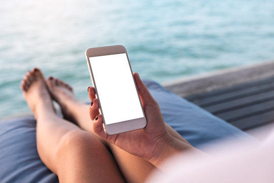 Mockup image of a woman using white mobile phone with blank desktop screen while sitting by the sea