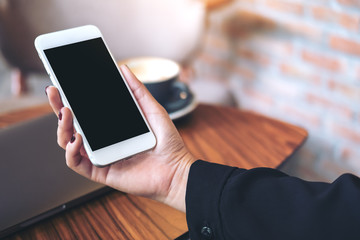 Mockup image of a woman's hand holding white mobile phone with blank black desktop screen and laptop on wooden table in cafe
