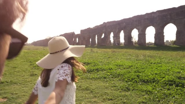Come with me. Young beautiful girl with hat leading man boyfriend holding hands toward roman aqueduct arches in parco degli acquedotti park ruins in rome romantic lovely happy couple slow motion