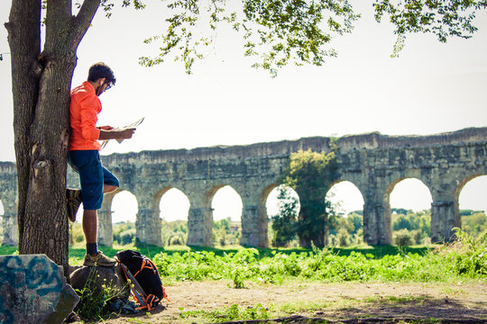 Backpacker standing on a rock under a tree reading a map in front of ancient roman aqueduct. Young attractive athletic man with orange sportswear and backpack in acquedotti park in Rome