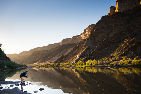 A hiker collecting water from a river reflecting the golden light of sunrise in the Fish River Canyon with high mountains reflected in  the calm water