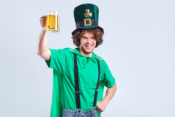 A young curly-haired man in the suit of St. Patrick with a mug of beer in his hand on a gray background.