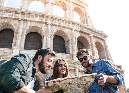 Three happy young friends tourists at Colosseum in Rome reading map guide for directions on sunny day. Ground shot with lens flare.