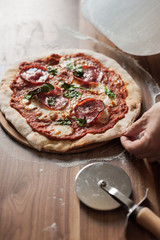 The hand takes a piece of finished pizza with tomato paste, salami, cheese and basil. Next to the wooden table lies a knife for pizza. Vertical