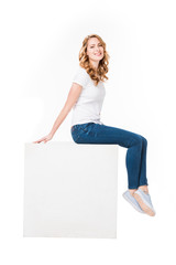 Fototapeta na wymiar cheerful woman sitting on empty white cube isolated on white