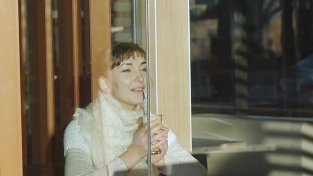 Young woman drinks coffee in cafe. Portrait of caucasian woman with lace wool scarf enjoying coffee aroma smiling and sipping latte through a straw behind a window.