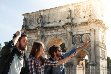 Three happy young friends tourists standing in front of Arch of Constantine in rome pointing directions. Lens flare.