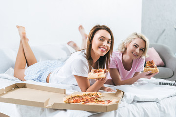 Obraz na płótnie Canvas smiling girls spending time together and eating pizza in bed