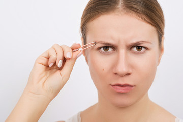 Woman plucking eyebrows with tweezers over grey background.