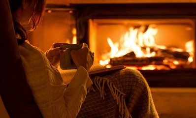 Young woman  sitting at home by the fireplace and reading a book. - obrazy, fototapety, plakaty