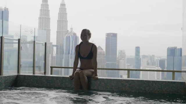 Pretty slim rich blonde woman in black bikini relaxing at the edge of roof top swimming pool or jakuzzi at resort or in hotel with view over city skyline. Success, vacation leisure activity concept.