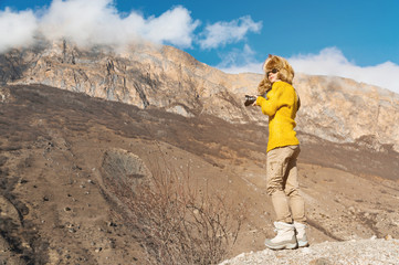 A girl photographer in sunglasses and a big fur hat and a yellow knitted sweater stands against the background of high rocks in the gorge with a dslr camera her in hand.