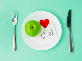 Ripe raw juicy green Apple, red heart, slip of paper with inscription diet on a white plate, fork, knife on a bright turquoise blue colour table, top view. Diet for heart health as a lifestyle.