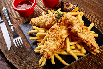 Black plate with fish and chips, mayo and ketchup on wooden background