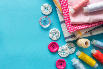 Multicolored threads, scissors, buttons, fabric and various sewing accessories on a blue background with copy space flat lay