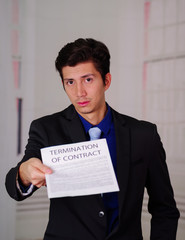 Close up of sad young businessman wearing a suit and holding a sheet of paper getting from the boss, with termination of contract text on it, in a blurred background