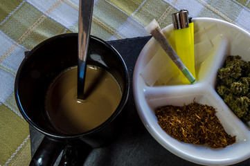 Materials for rolling joints: high quality marijuana buds, chopped tobacco, lighter and paper. Plate accompanied by a joint and a cup of coffee with milk.