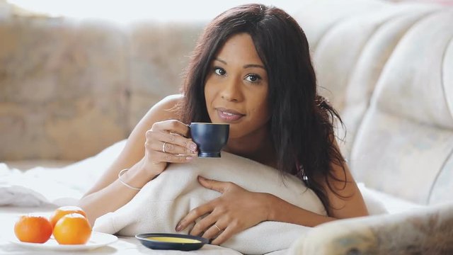 Brunette woman in cosily bed drinking cup of tea at home