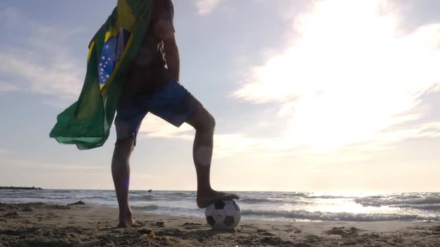 Young man wearing brazilian brazil national flag as super hero cape stands with foot on football on the sea shore sand looking at the ocean at the beach at sunset camera steadycam gimbal revolving